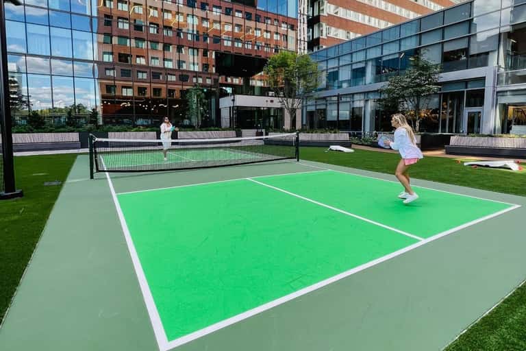 Outdoor pickleball court with green playing surface surrounded by manicured lawn and urban buildings