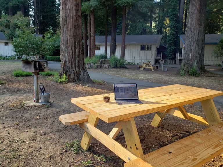 Wooden picnic table with laptop in forest campground setting among tall redwood trees and cabins