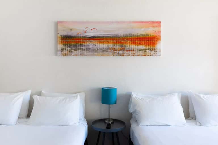 Two twin beds with white linens separated by teal bedside table and lamp beneath abstract landscape artwork