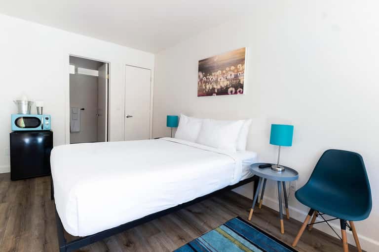 Modern hotel room with queen bed, white bedding, teal accent lamps, wooden floors, and retro microwave on black dresser