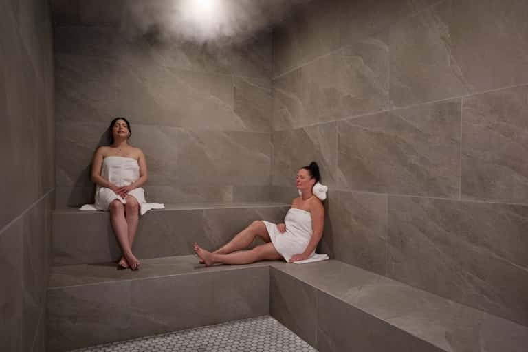 Two women relaxing on stone benches in a spa steam room with gray tile walls