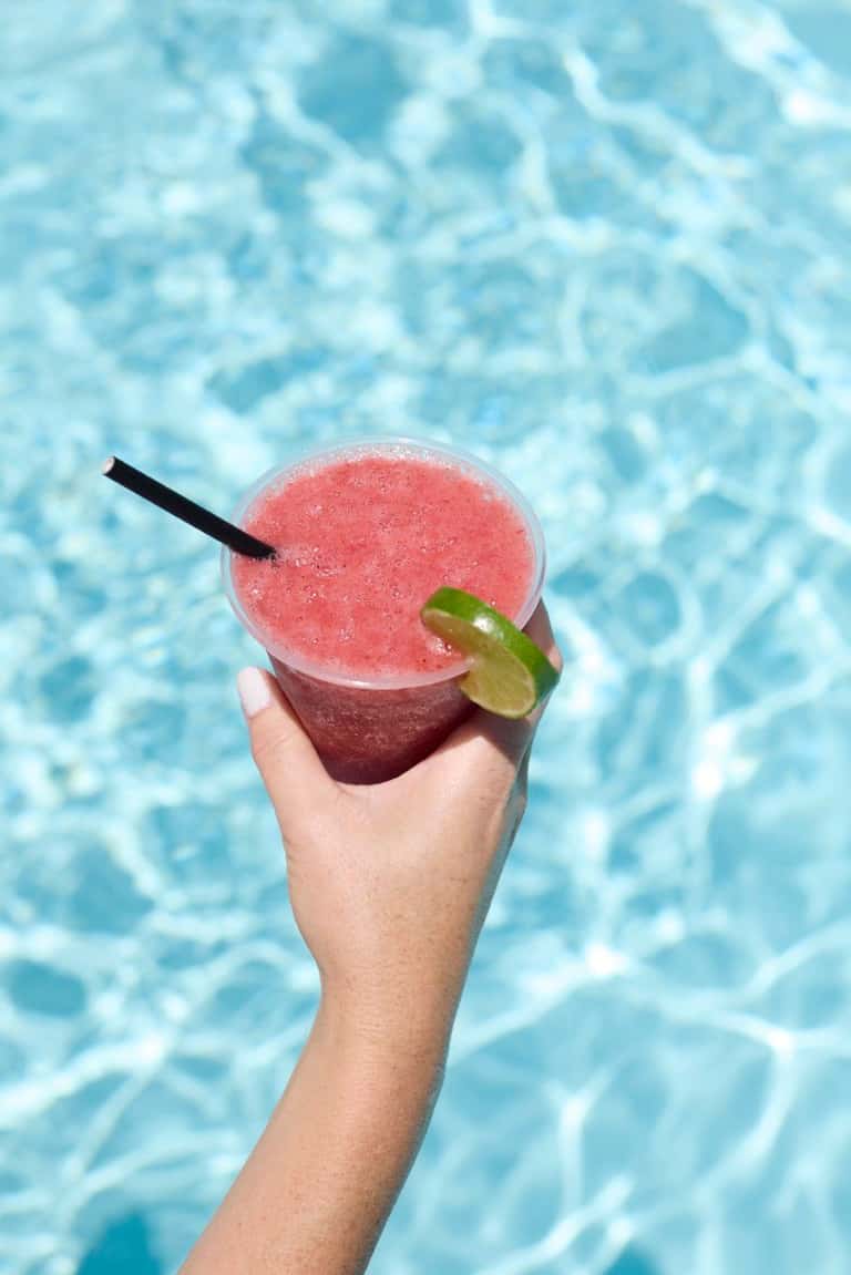 Hand holding pink frozen cocktail with lime garnish over pool water background