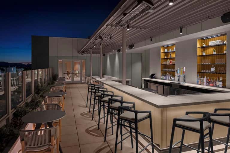 Rooftop bar at night with wooden shelving, white counter, and black bar seating