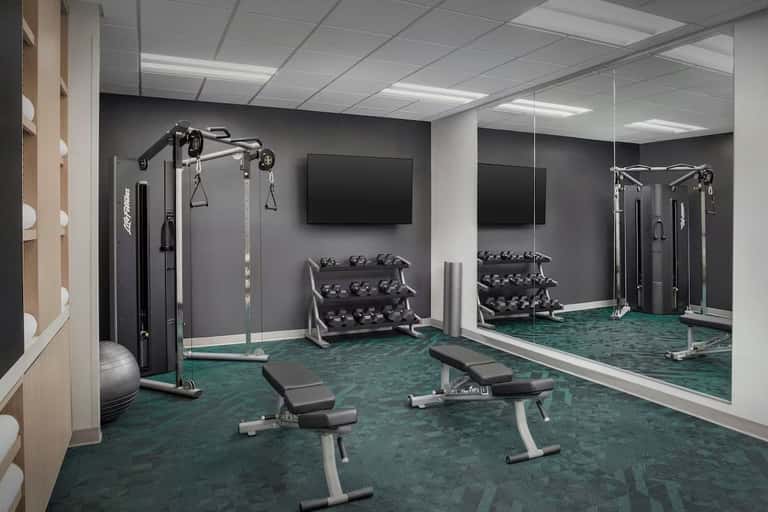 Modern fitness center with weight racks, cable machines, benches, mirrors, and TV on teal carpeting