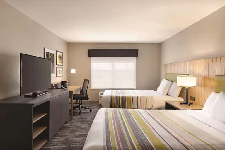 Modern hotel room with two queen beds, striped bedding, desk, and window with dark shade