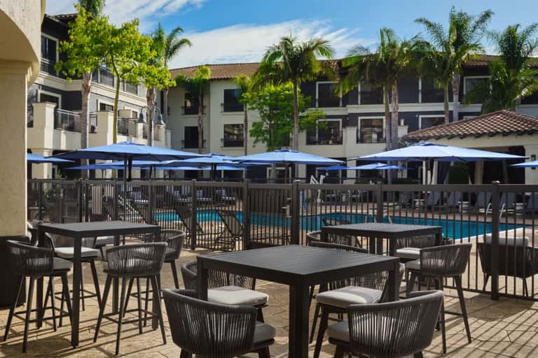 Resort poolside patio with blue umbrellas, black rope chairs, and dining tables overlooking the swimming pool