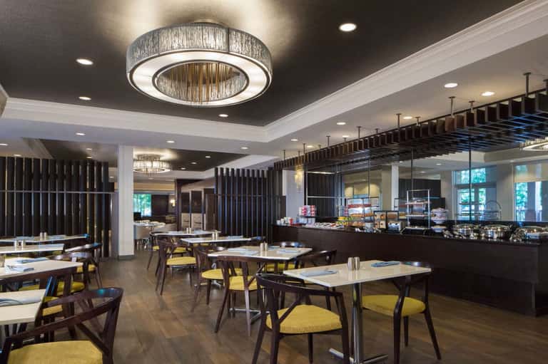 Hotel restaurant and breakfast area with dining tables, yellow chairs, serving counter, and contemporary lighting