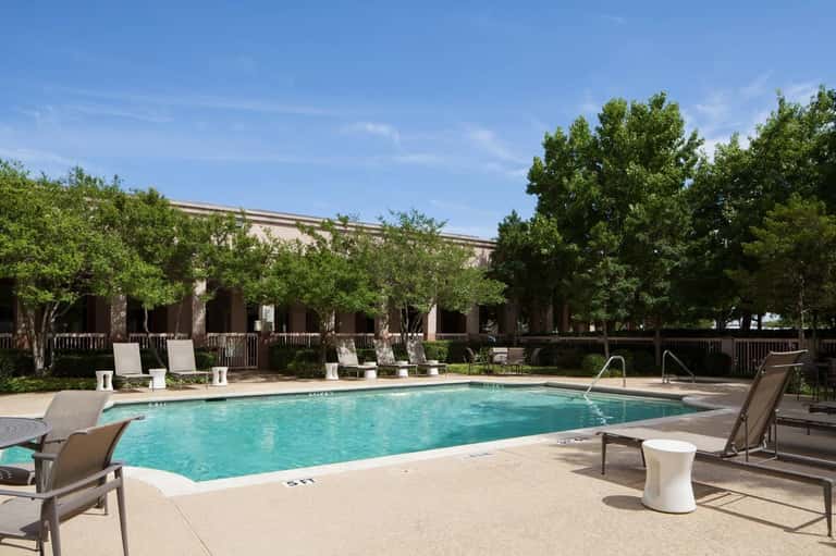 Rectangular resort pool with turquoise water, lounge chairs, and brick building with white railings surrounded by mature trees