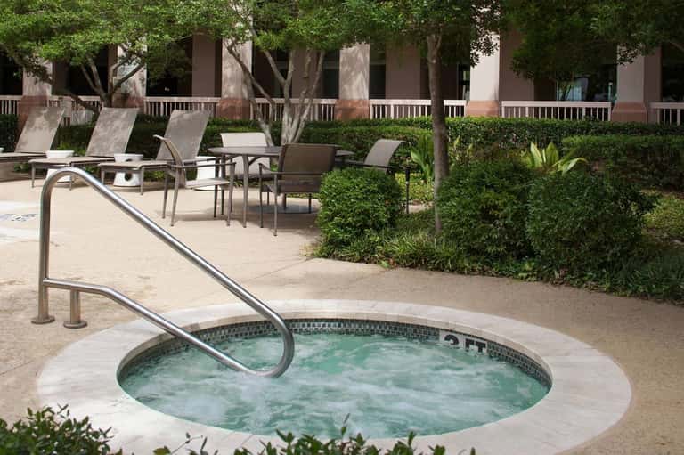 Circular hot tub with metal handrails on concrete patio, lounge chairs, brick building and landscaping in background