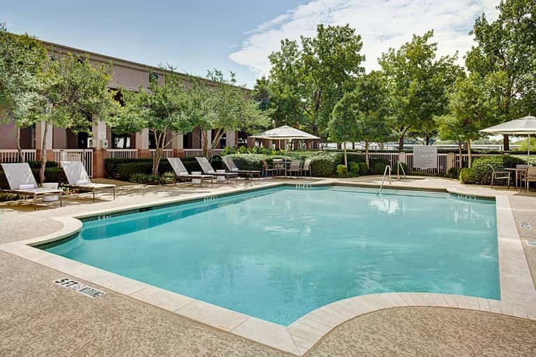 Curved resort swimming pool with turquoise water, white umbrellas, lounge chairs, brick building, and mature trees lining the deck