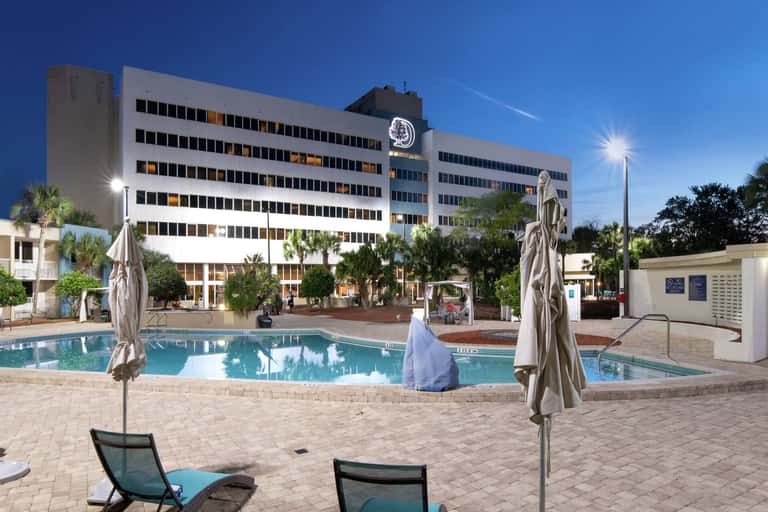 Hotel outdoor pool area at dusk with turquoise water, umbrellas, lounge chairs, multi-story building backdrop, and palm trees
