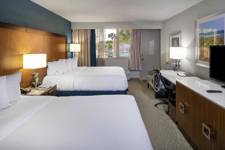 Modern hotel guest room with two queen beds, wood headboard, teal accent wall, and glass sliding door to balcony