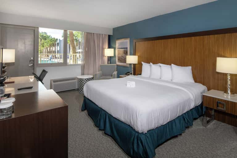 Modern hotel bedroom with wood headboard, blue accent wall, desk, and ocean view through large windows