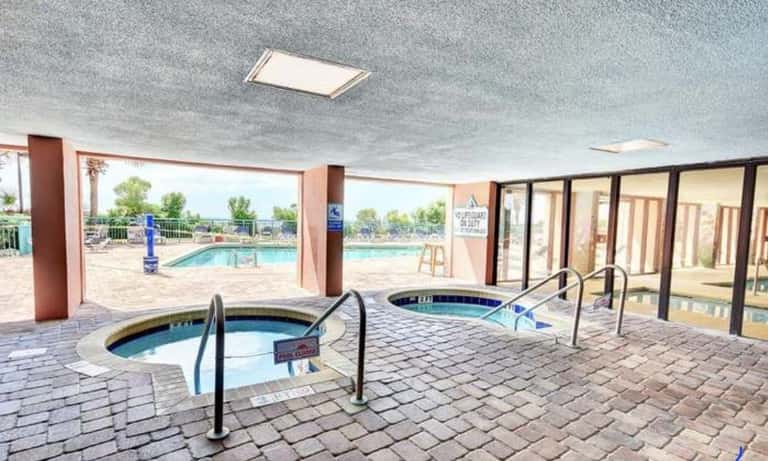 Indoor spa area with two hot tubs, columns, large windows, outdoor pool visible, and glass patio doors to beach or grounds
