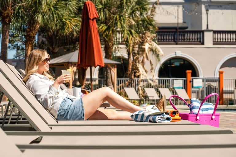 Woman lounging by resort pool with red umbrella, palm trees, and modern building architecture
