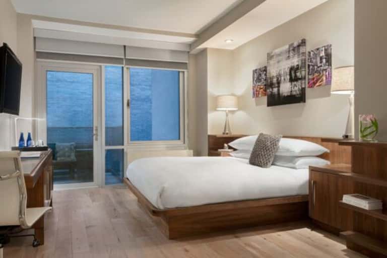 Modern hotel bedroom with king bed, wood furnishings, and floor-to-ceiling windows overlooking city skyline at dusk