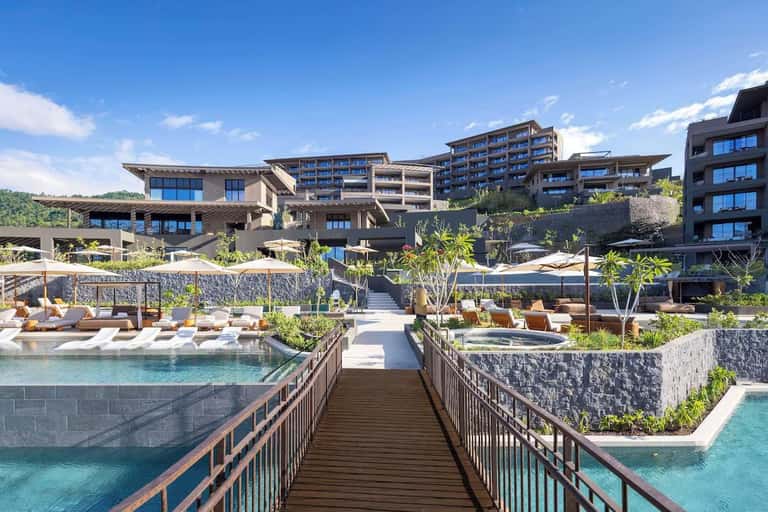 Modern resort complex with tiered architecture, multiple pools, wooden walkway, and lush tropical landscaping