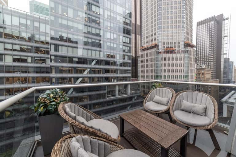 High-rise hotel balcony with two woven chairs, wood table, city skyline views, and glass railing
