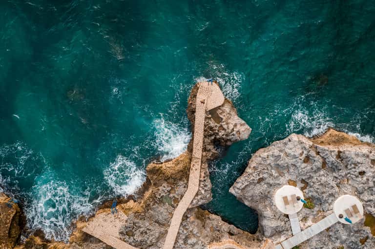 Aerial view of stone pathway leading to sea platforms with turquoise ocean and white umbrellas below