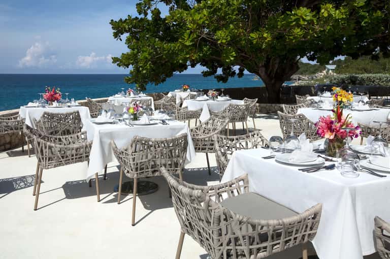Beachfront dining setup with white tables, woven chairs, and tropical flowers by the sea