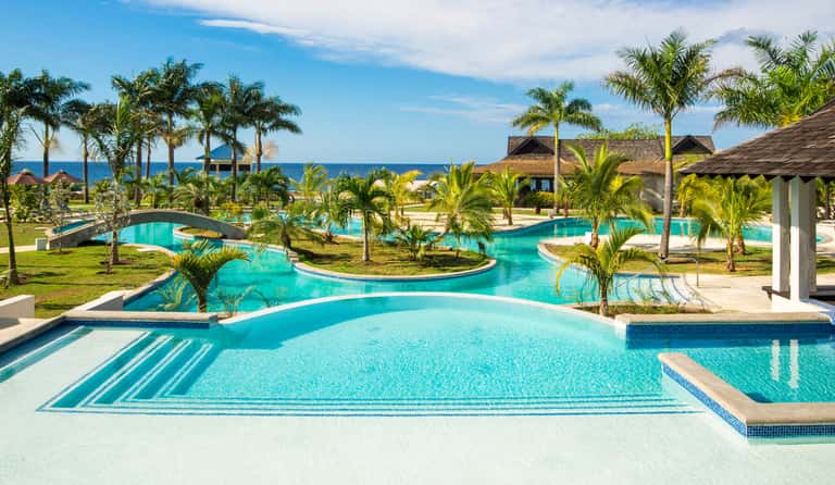 Resort pool complex with winding waterways, palm trees, beach access, and thatched structures