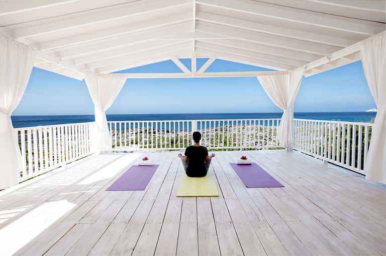 Oceanfront yoga studio with white ceiling beams, white railings, flowing curtains, and person meditating on purple mat