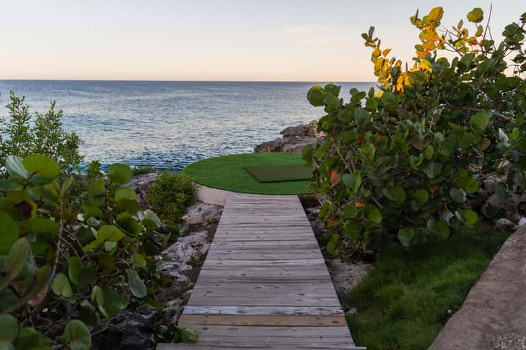 Wooden boardwalk path through tropical vegetation leading to ocean and manicured lawn area at dusk