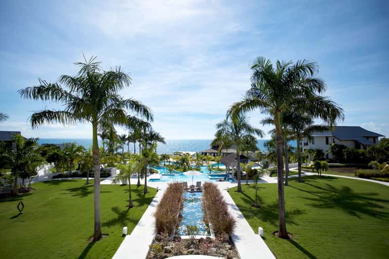 Luxury resort grounds with palm-lined pathway, rectangular pool, manicured lawn, and ocean view in background