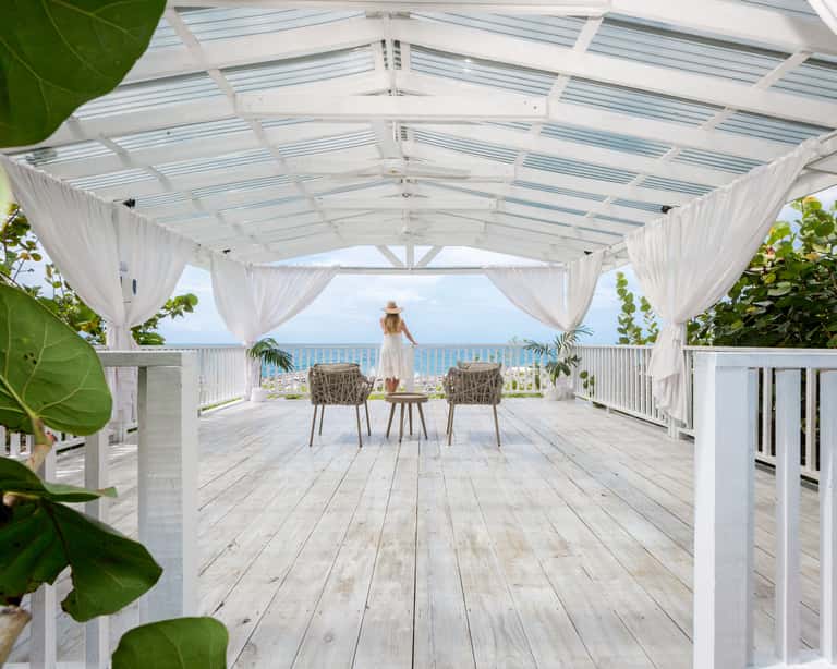 White pergola with sheer curtains on wooden deck overlooking calm blue ocean and coastline