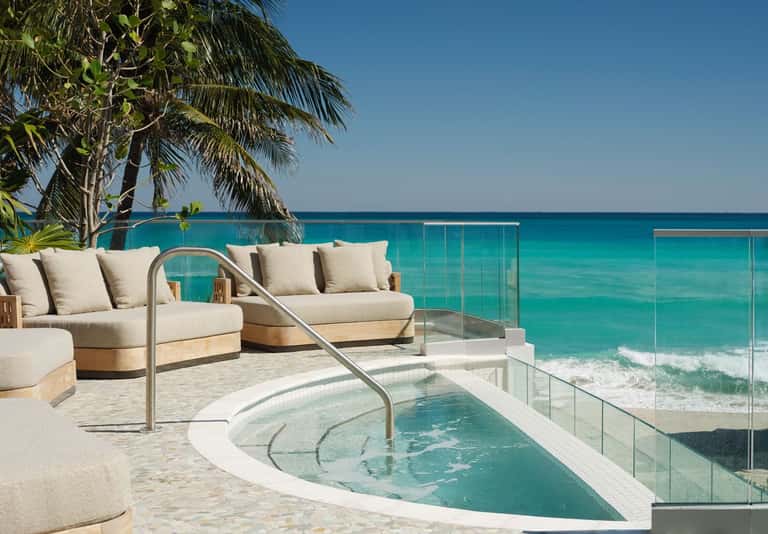 Beachfront infinity pool overlooking turquoise ocean with cream loungers, palm trees, and glass railing on pristine sand