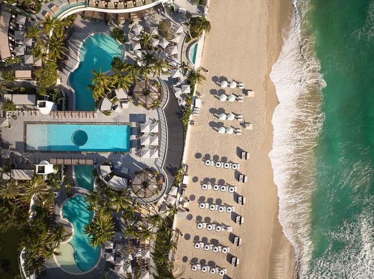 Aerial view of beachfront resort with pools, lounge chairs, palm trees, sand, and turquoise ocean