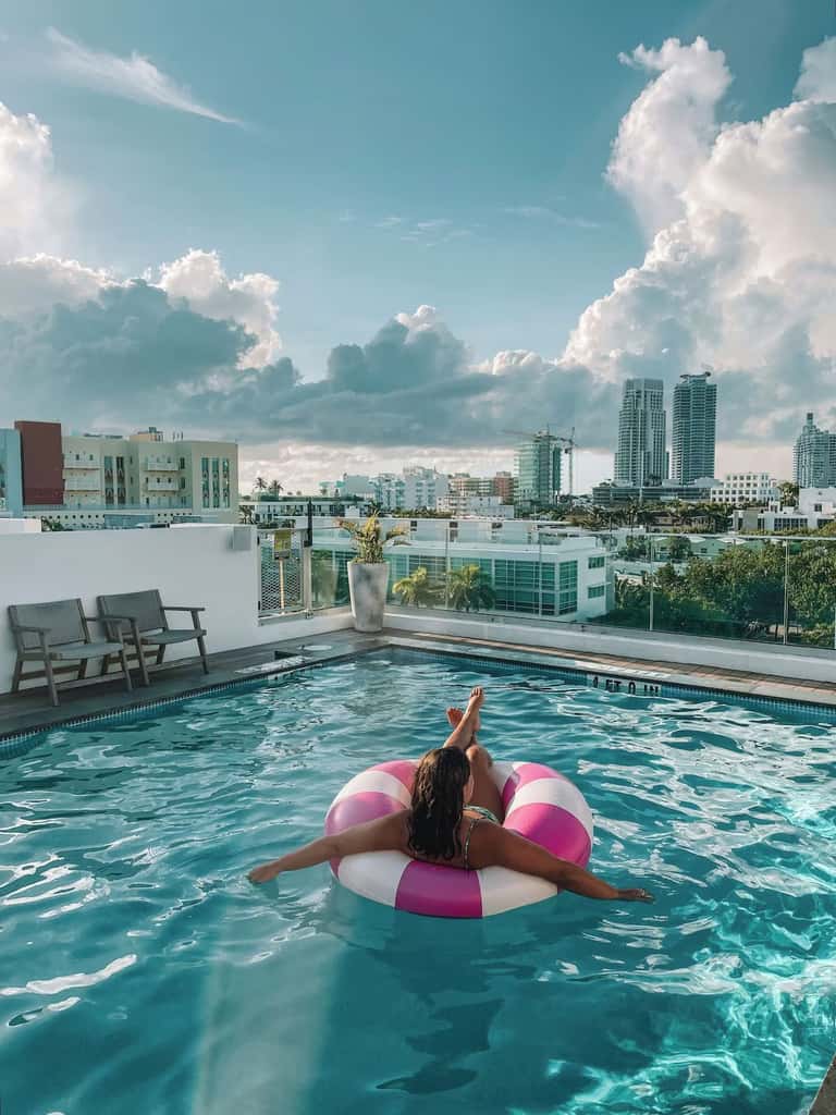 Woman on pink and white float in rooftop pool with city skyline and dramatic clouds