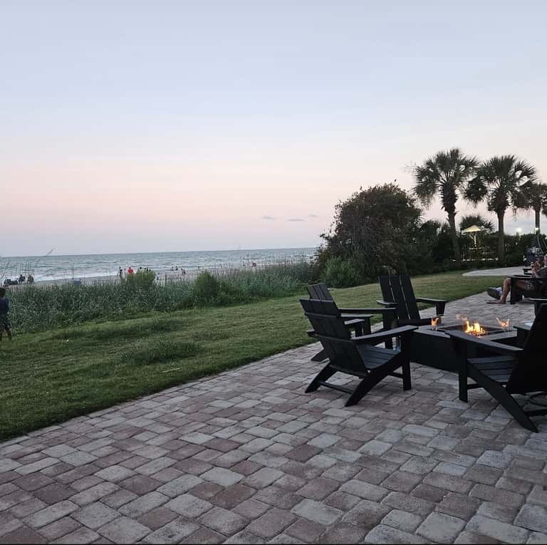 Beachfront fire pit at dusk with black Adirondack chairs, ocean view, palm trees, and coastal landscape