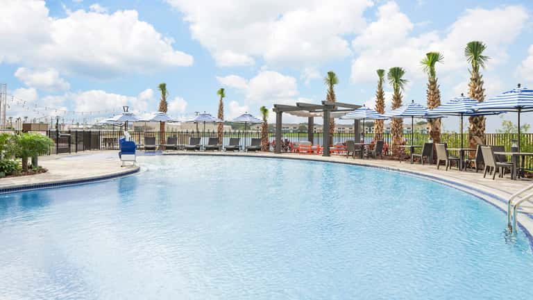 Resort pool during daytime with palm trees, blue umbrellas, lounge chairs, and scenic landscaping