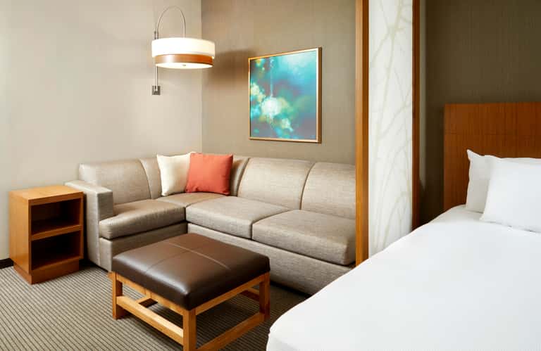 Modern hotel suite with beige sectional sofa, brown ottoman, wood nightstand, and ocean-themed artwork above bed