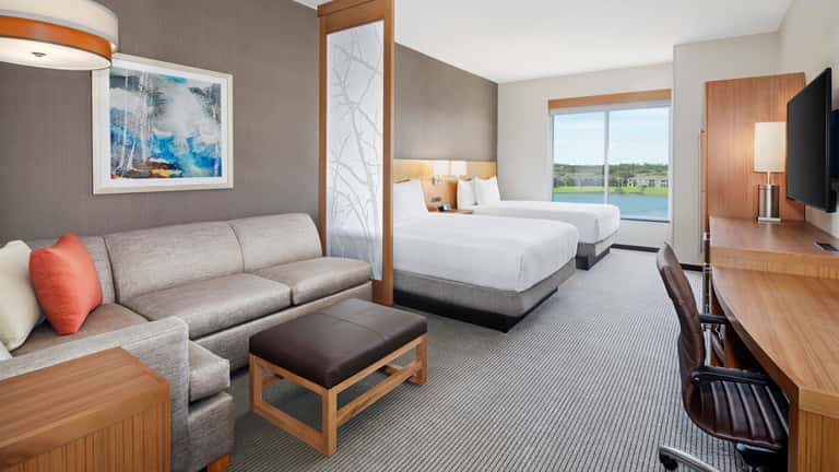Spacious hotel suite with two queen beds, gray sofa, wood furnishings, large windows overlooking golf course