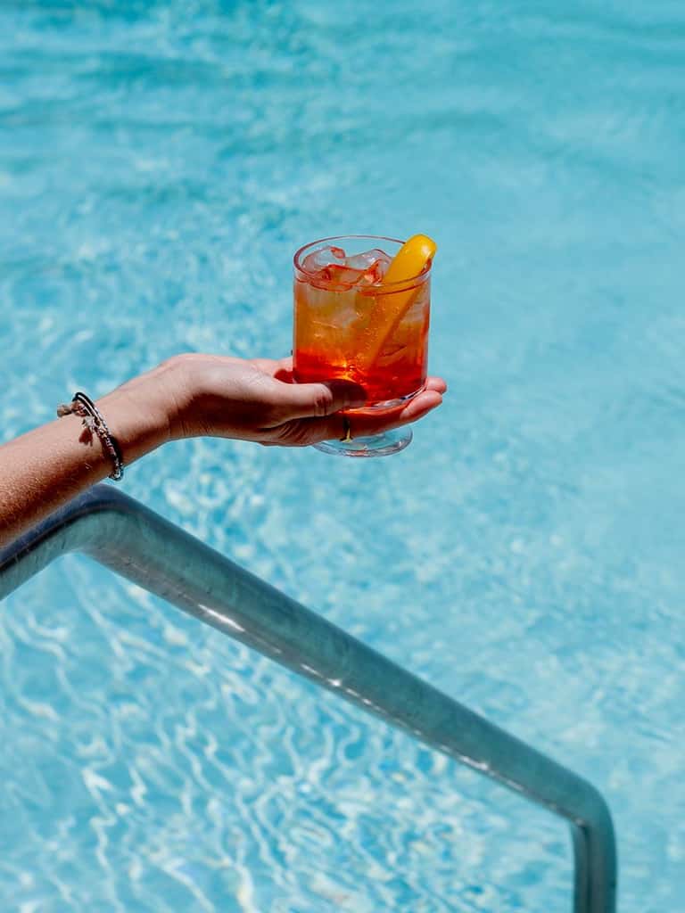 Hand holding a red cocktail with ice and lemon garnish over a swimming pool