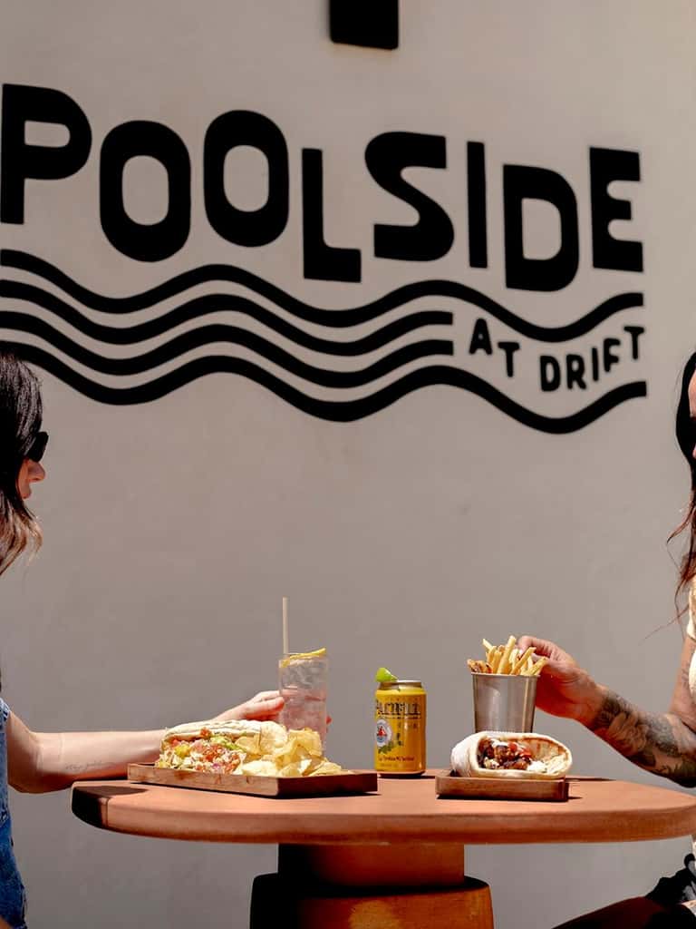 Two people sharing food at a round table with 'Poolside at Drift' signage behind them