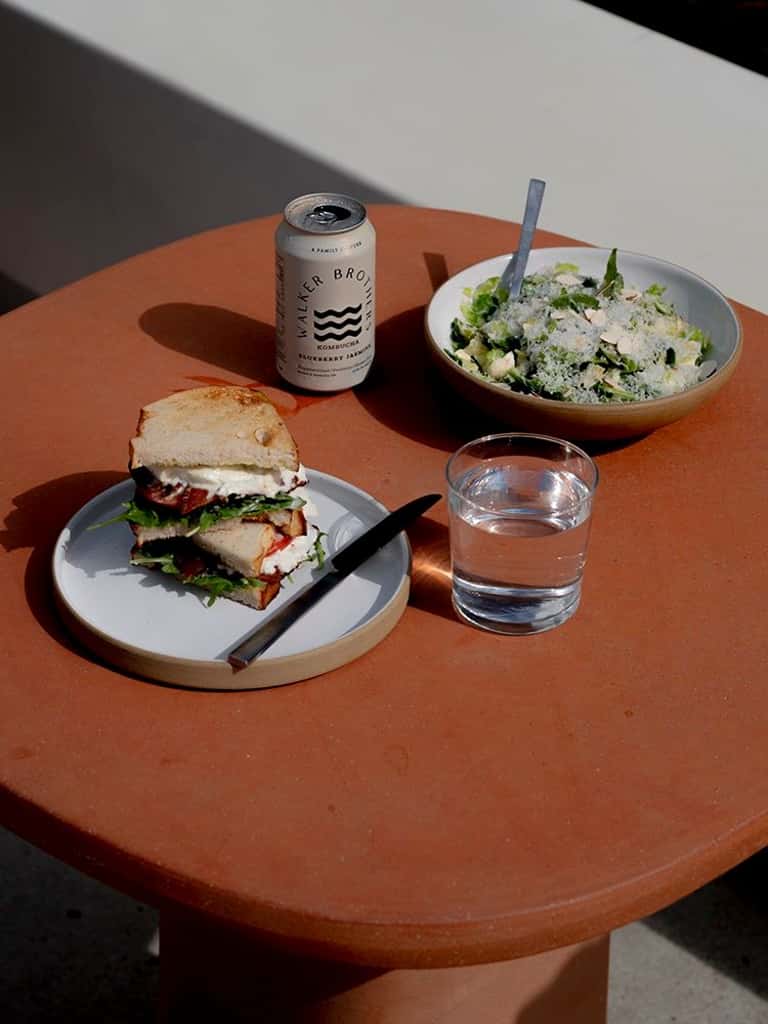 Outdoor dining table with sandwich, salad bowl, beverage can, and water glass on terracotta surface