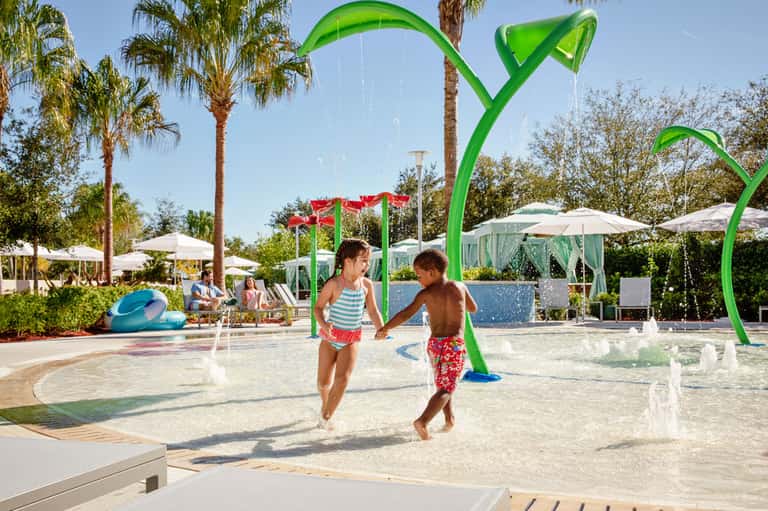Children playing in shallow splash pad with green water features and palm trees at resort
