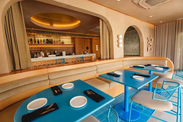 Modern restaurant dining area with blue tables, curved beige banquette seating, and illuminated bar counter in background