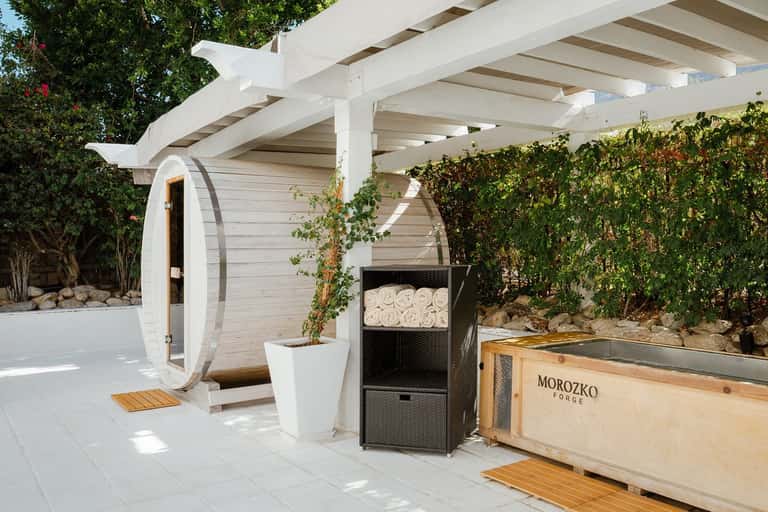 Modern white pergola structure with rolled towels storage and potted plants near wooden hot tub in a minimalist spa area