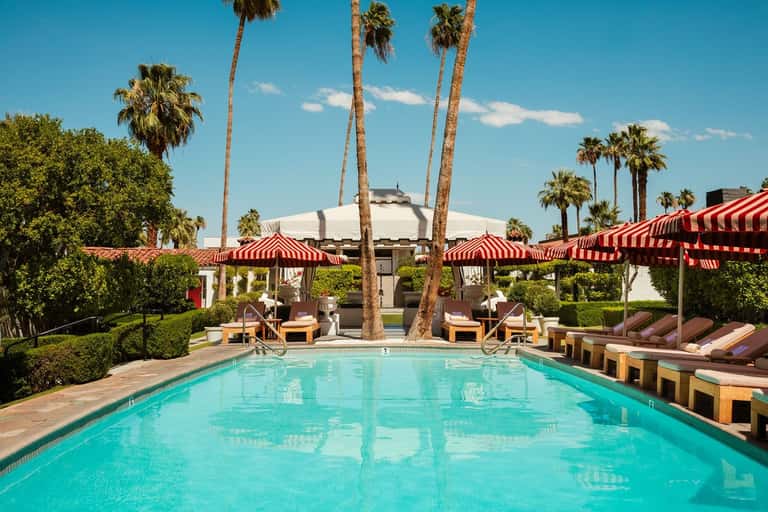 Olympic-sized pool with red striped umbrellas, white cabanas, palm trees, and modern resort building architecture