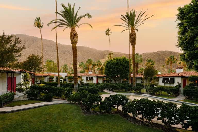 Desert resort grounds at sunset with white bungalows, palm trees, and mountains reflected in evening light