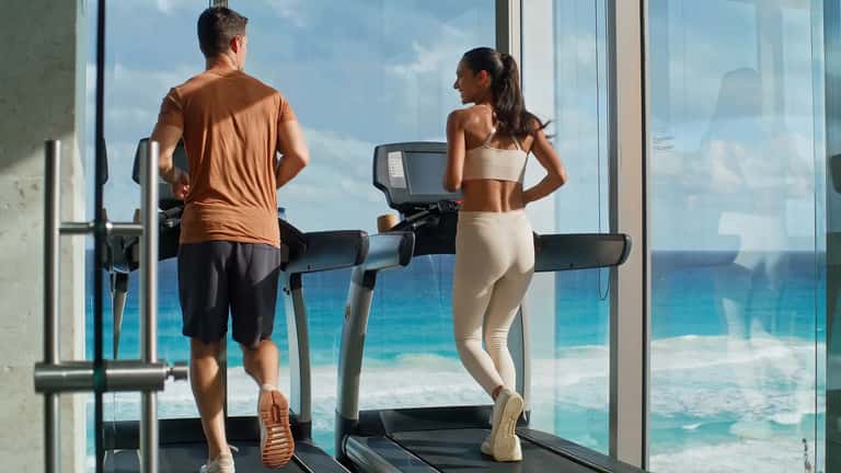 Two people exercising on treadmills with floor-to-ceiling windows overlooking beach and ocean