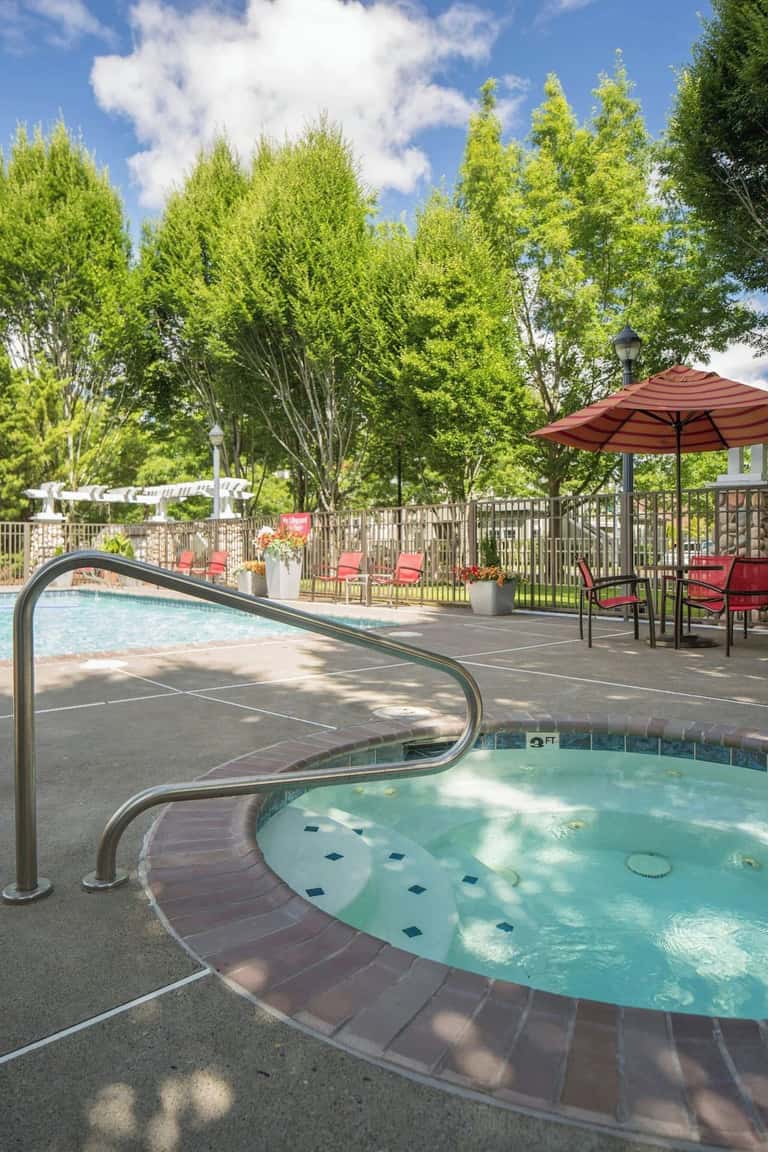 Spa-like hot tub area surrounded by tall trees, lounge chairs, red umbrella, and serene outdoor setting