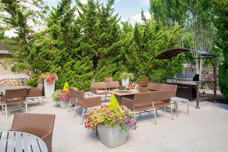 Outdoor patio seating with wicker chairs, vibrant flower planters, grill station, and green tree backdrop