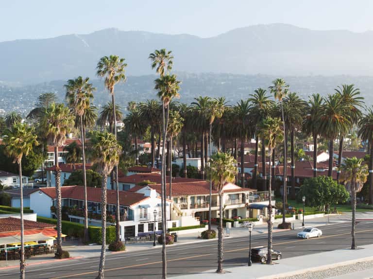 Palm tree-lined street with mountain views, residential buildings with Spanish-style tile roofs, and hillside landscape