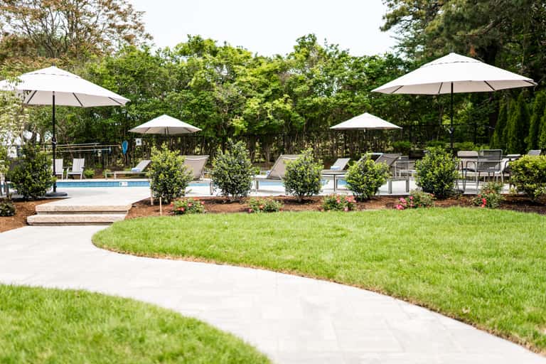 Poolside patio with white umbrellas, lounge chairs, manicured lawn, and surrounding trees