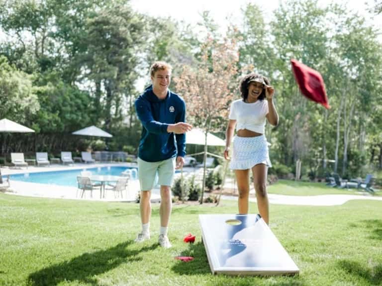Two guests playing cornhole on grass near resort pool with trees and white umbrellas